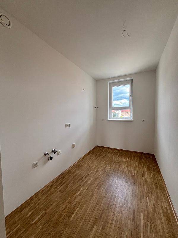 Leerer Raum mit Parkettboden und Anschlüssen für eine Küche, hell durch ein Fenster.