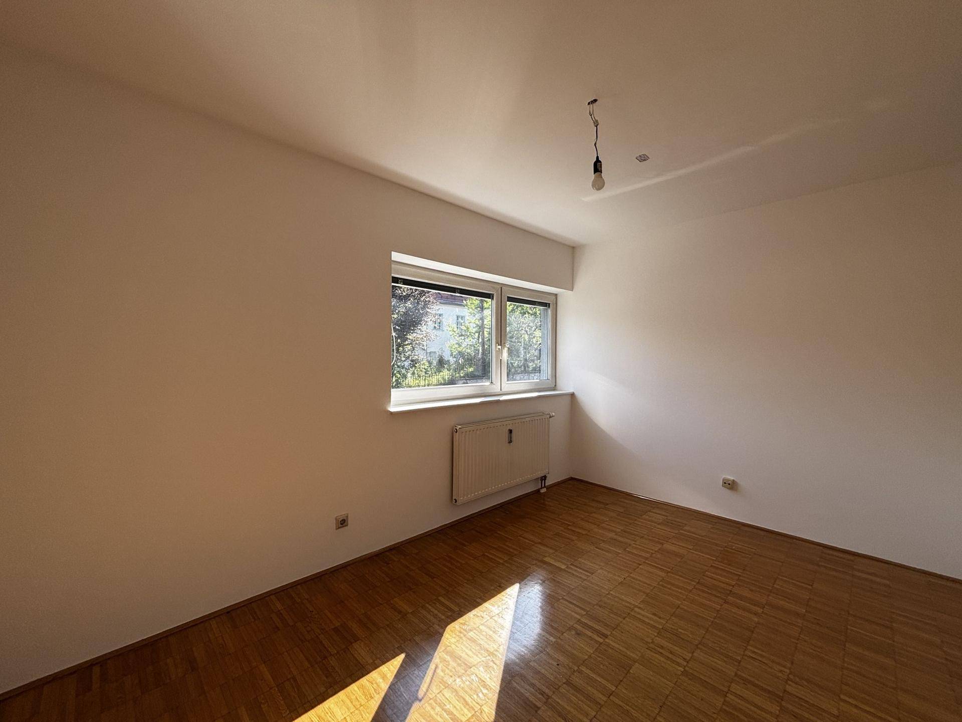 Helles Schlafzimmer mit Parkettboden, weißen Wänden und einem Fenster mit Blick ins Grüne.