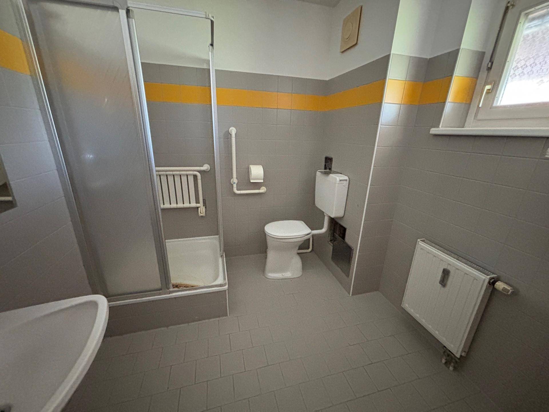 Badezimmer mit grauen und gelben Fliesen, Duschkabine, WC und Fenster für Tageslicht.