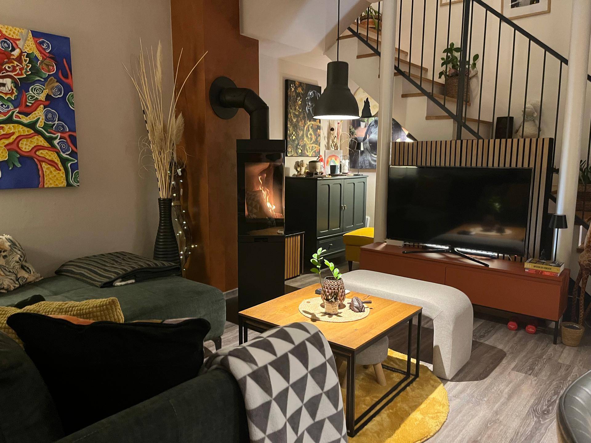 Gemütliches Wohnzimmer mit modernem Holzofen, Sofa und Fernseher, Treppe im Hintergrund.