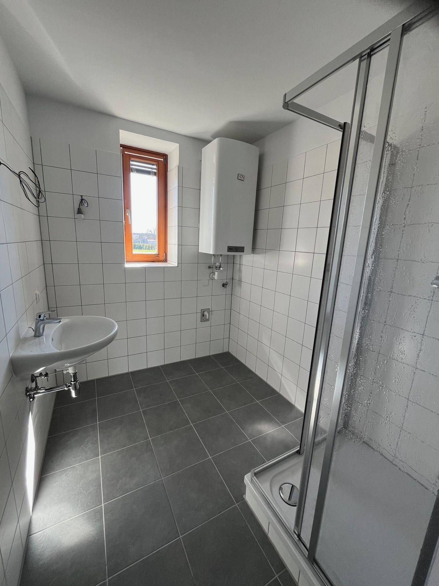 Badezimmer mit Duschkabine, Waschbecken, Fenster und grauen Bodenfliesen, hell und praktisch gestaltet.