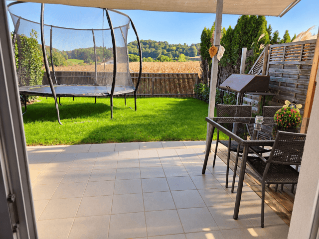 Geräumige Terrasse mit Gartenmöbeln, Grillplatz und Trampolin, umgeben von grüner Landschaft.