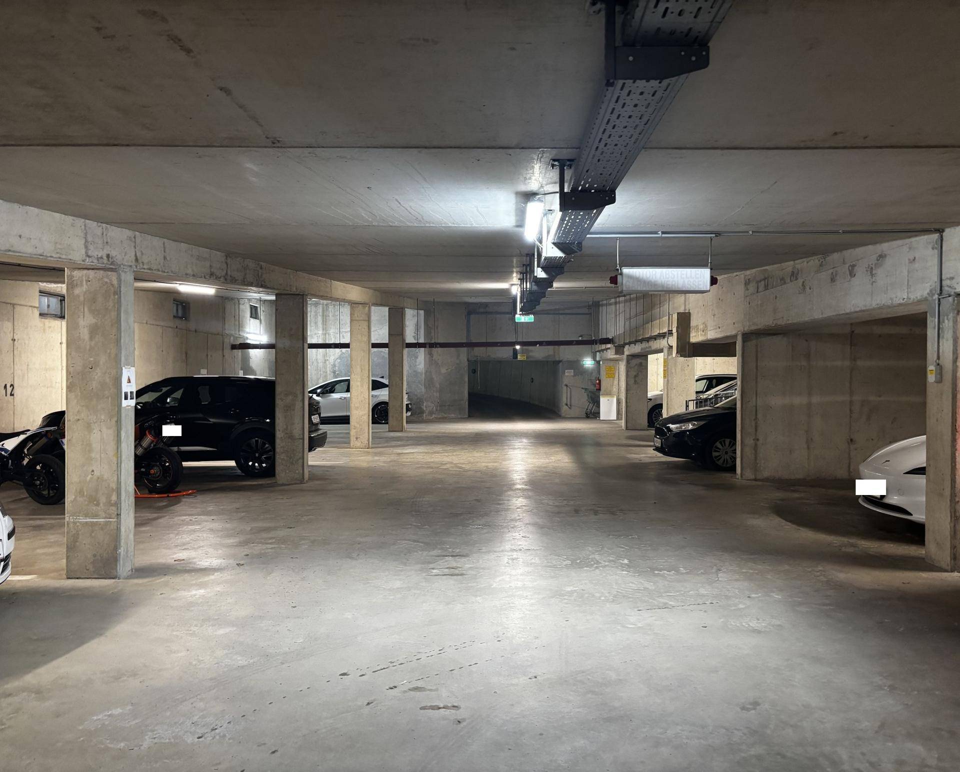 Geräumige Tiefgarage mit vielen Stellplätzen, Betonpfeilern und sichtbaren Kabeltrassen an der Decke.