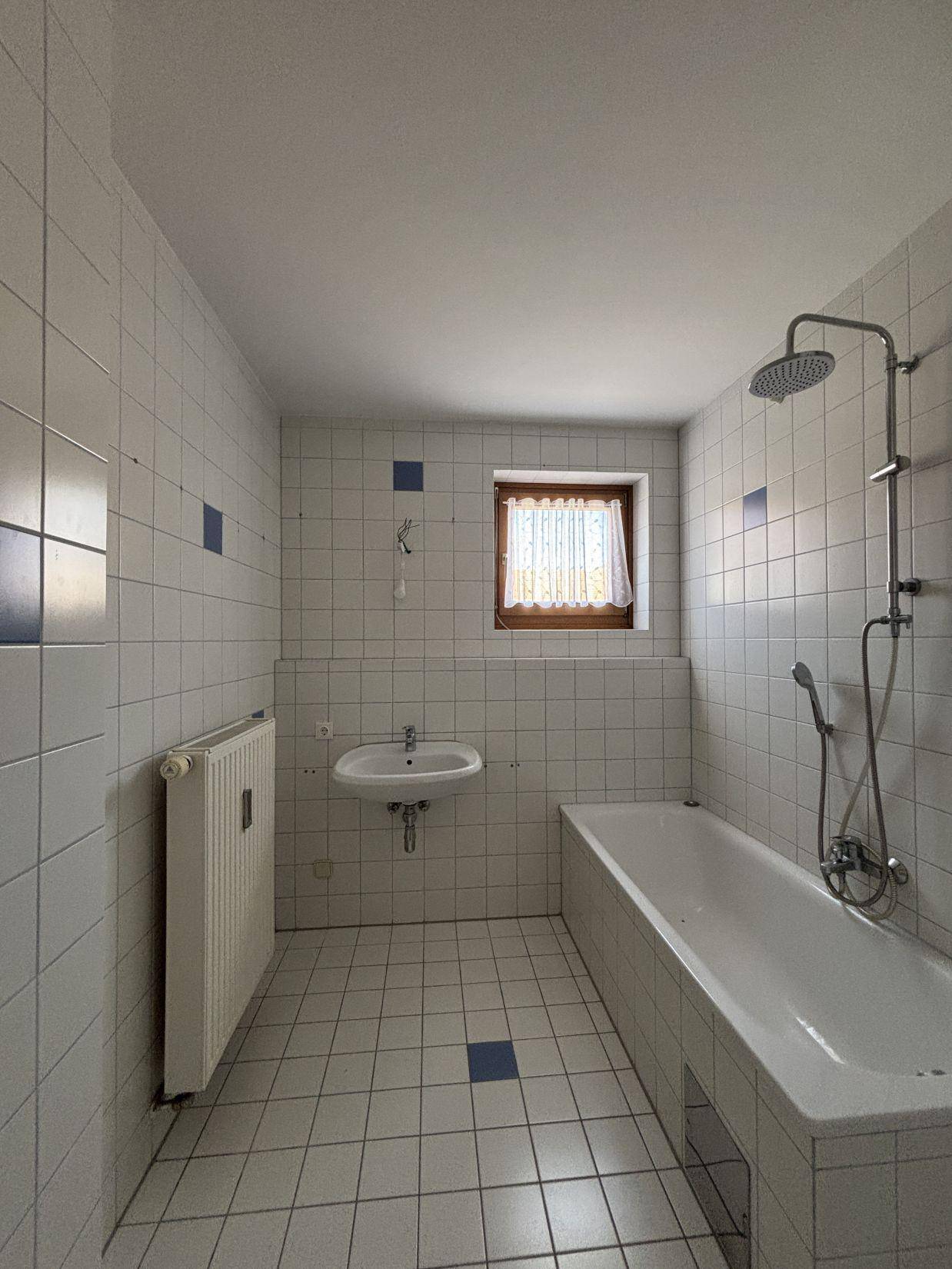 Badezimmer mit Badewanne, Duschvorrichtung, Waschbecken und Fenster, komplett gefliest.