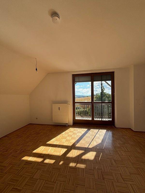 Helles Wohnzimmer mit Parkettboden, Dachschräge und großem Balkonzugang, der viel Sonnenlicht hereinlässt.