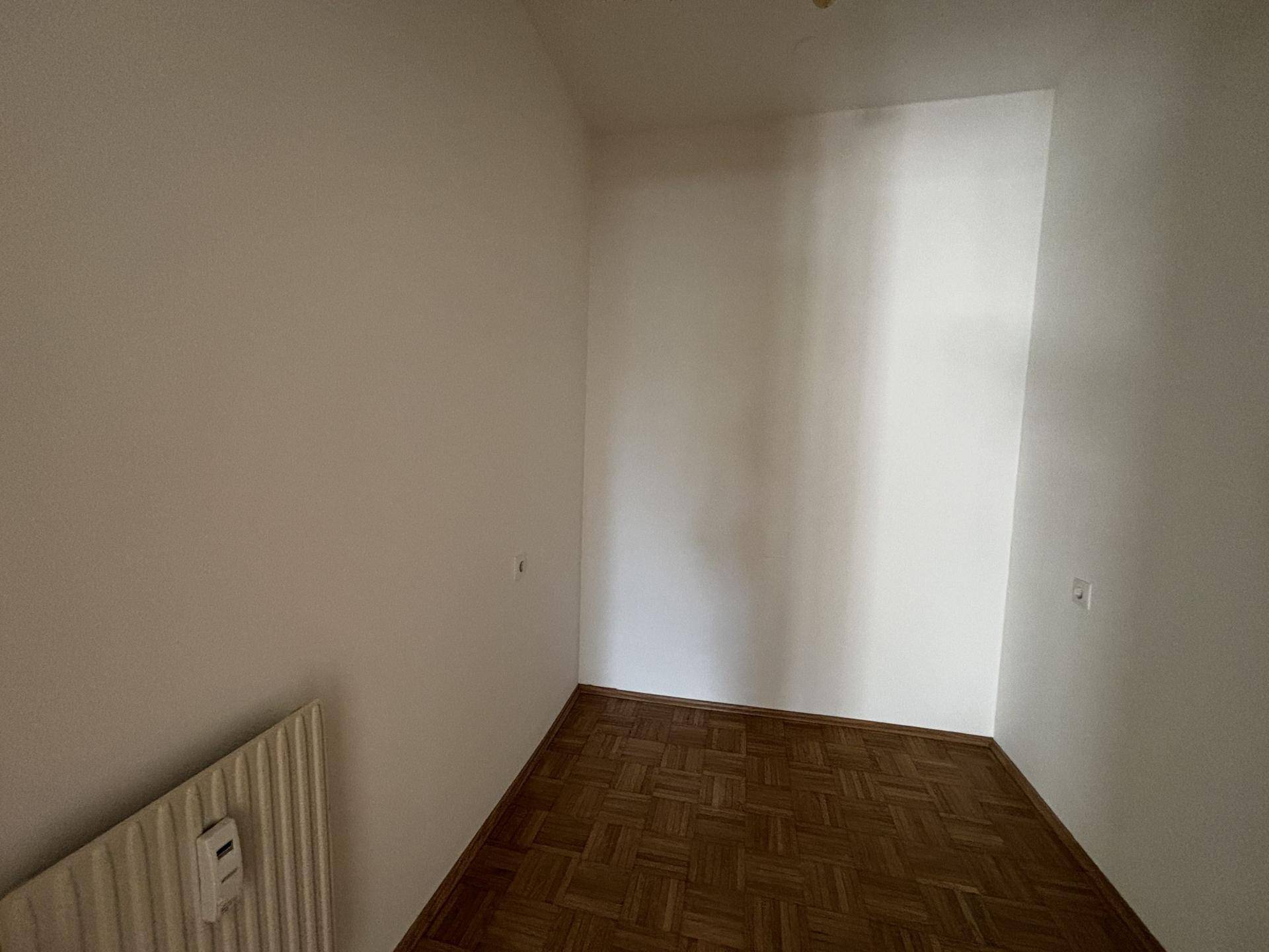 Kleiner leerer Raum mit Parkettboden und einem Heizkörper an der Wand, vielseitig nutzbar.