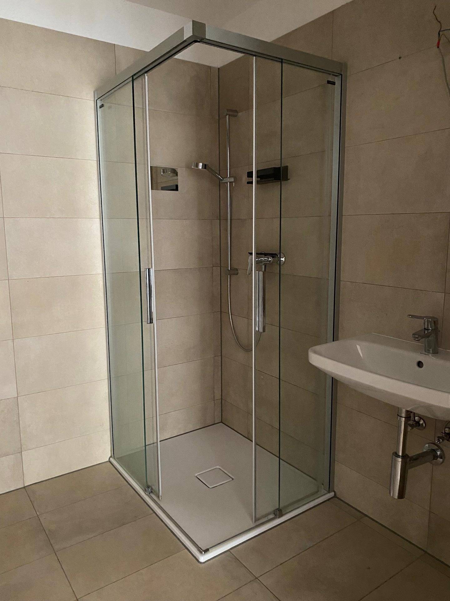 Modernes Badezimmer mit Duschkabine aus Glas, wandmontiertem Waschbecken und beigefarbenen Fliesen.