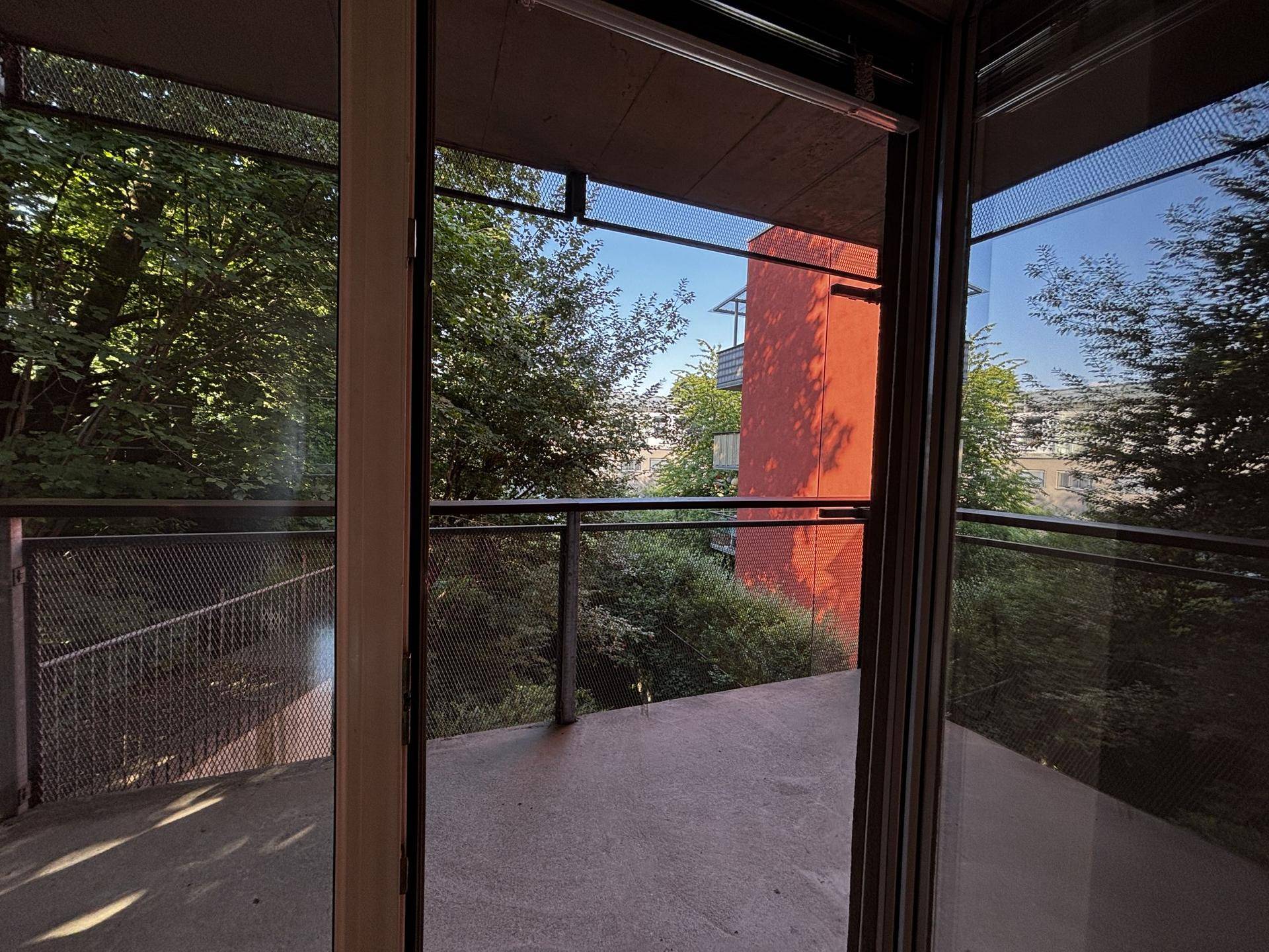 Blick vom Balkon ins Grüne mit Bäumen und einem weiteren Gebäude in der Ferne.