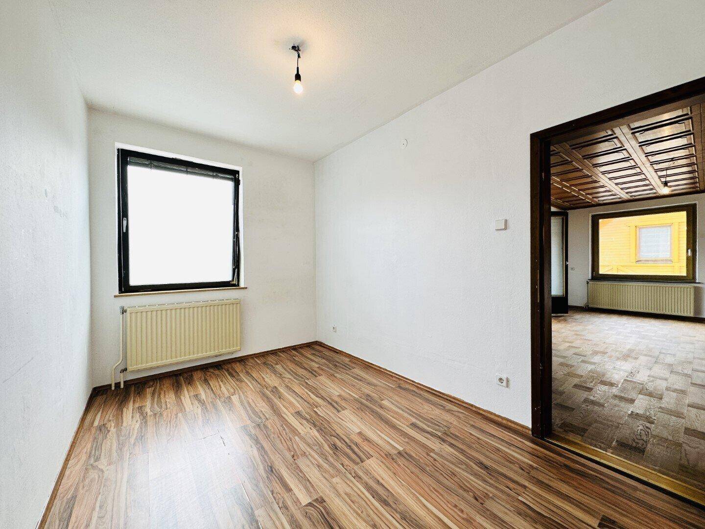 Leeres Zimmer mit Laminatboden und Fenster, verbunden mit dem Hauptwohnbereich.