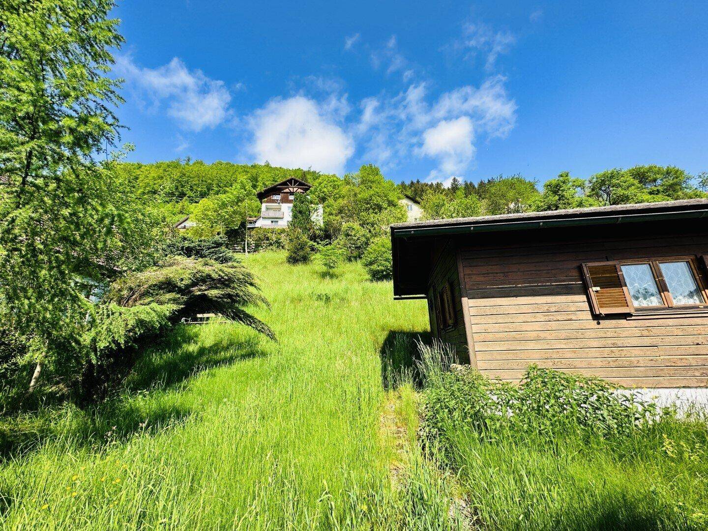 Kleines Holzgebäude in einer idyllischen, sonnigen Wiesenlandschaft mit bewaldeten Hügeln und Häusern im Hintergrund.