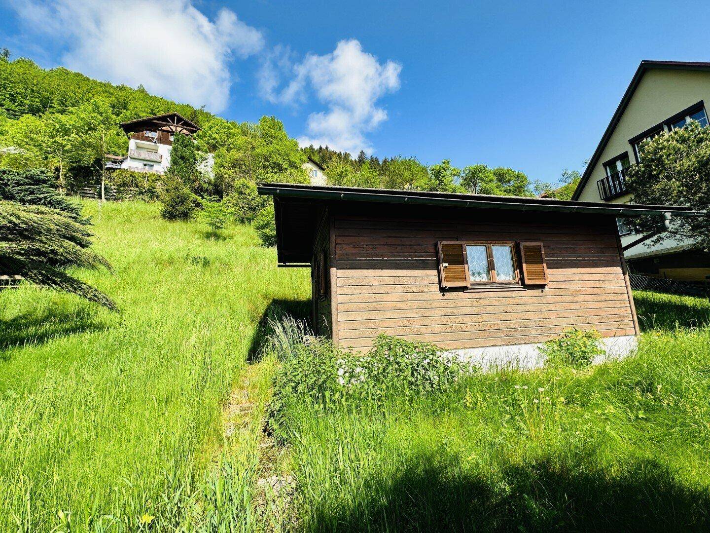 Rustikales Holzhaus eingebettet in eine sonnige, grüne Hügellandschaft mit weiteren Häusern im Hintergrund.