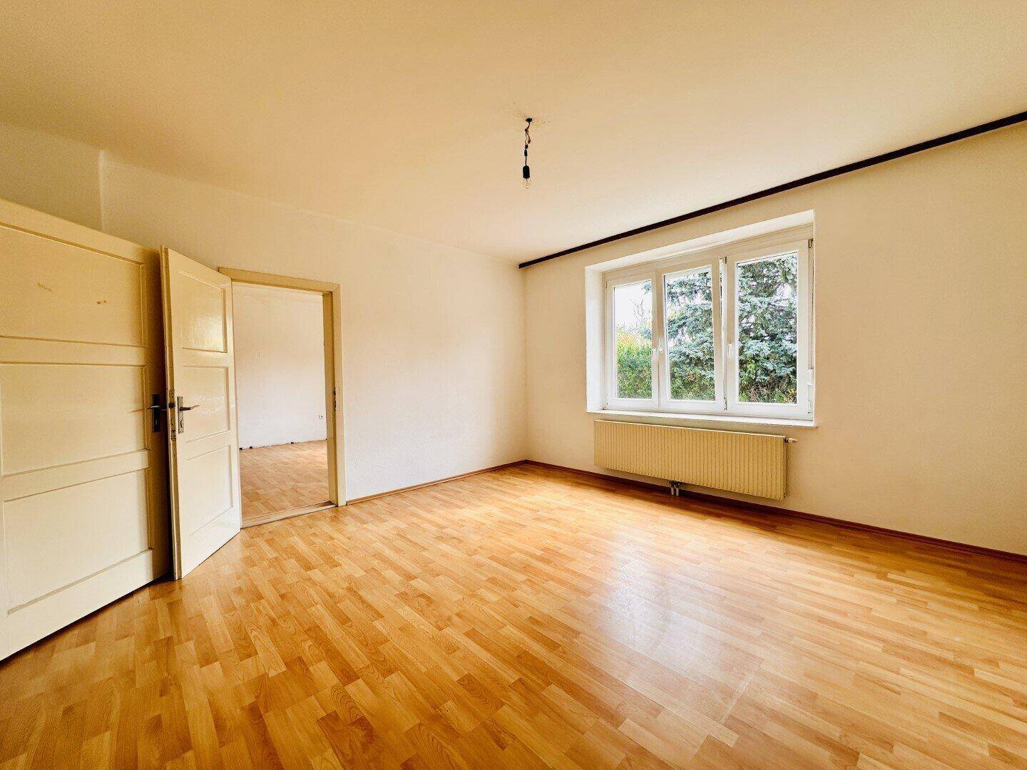 Helles, leeres Zimmer mit Holzparkettboden, großen Fenstern und Zugang zu einem weiteren Raum.