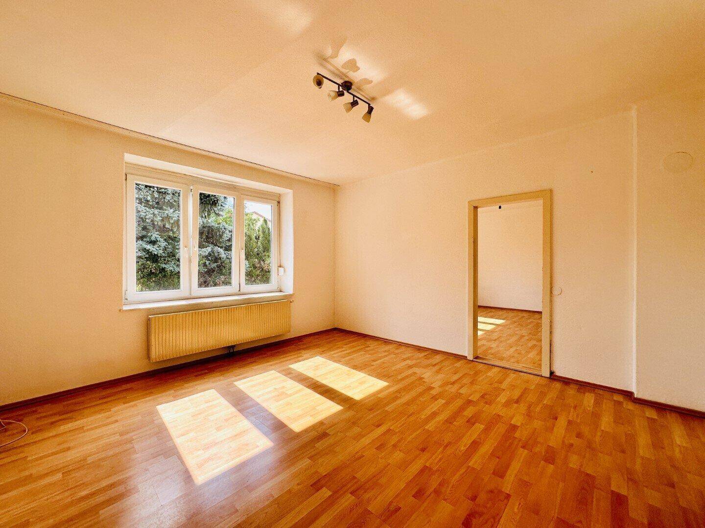 Sonnendurchflutetes Zimmer mit Holzparkettboden, großen Fenstern und einem angenehmen Blick ins Grüne.