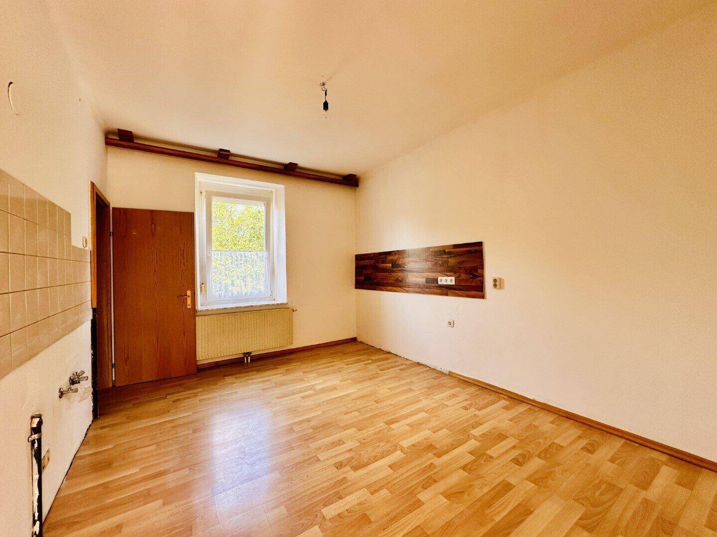 Leere Küche mit Holzparkettboden, gefliestem Bereich und einem Fenster mit Blick nach draußen.