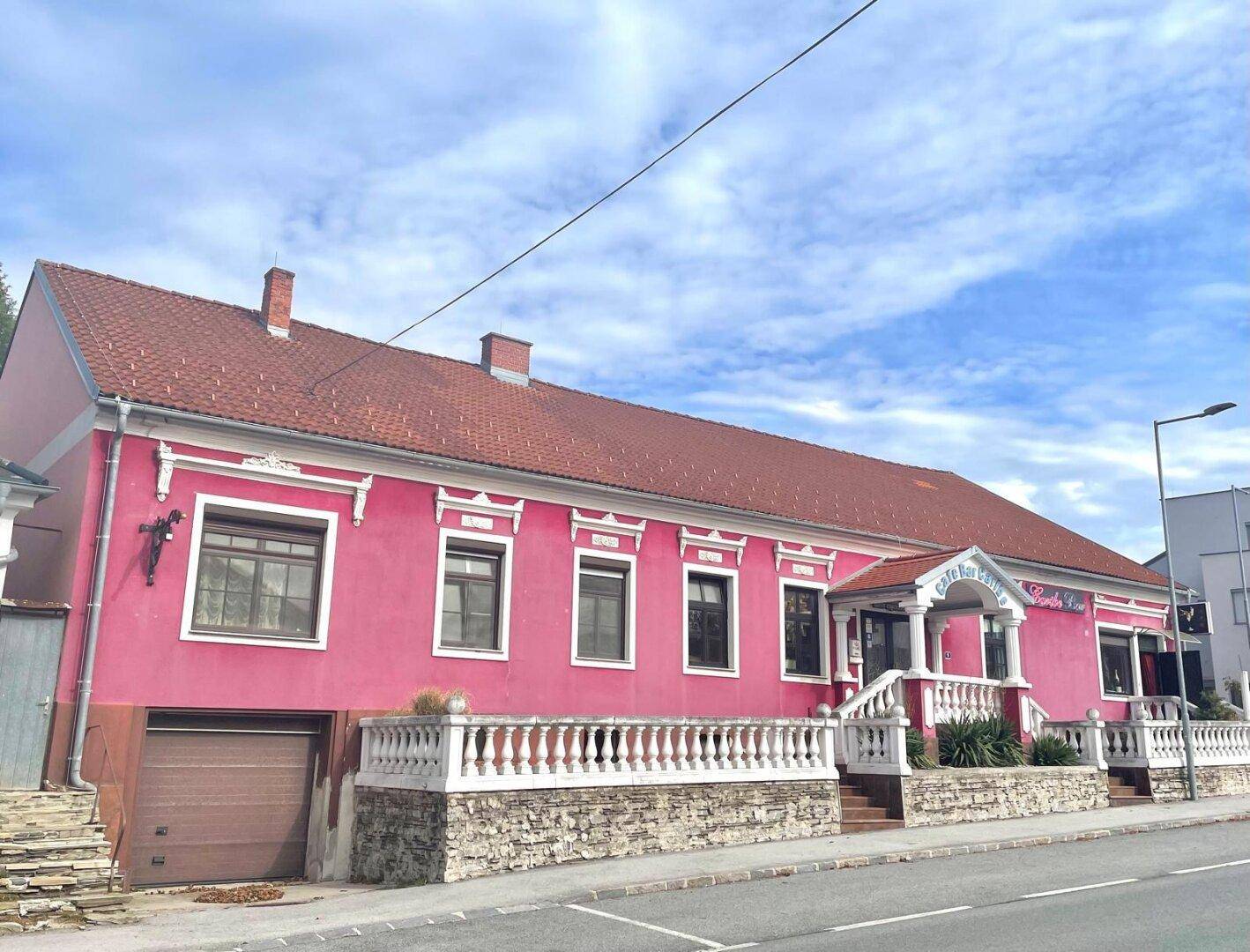 Gebäude mit auffälliger pinker Fassade, rotem Ziegeldach und dekorativen weißen Elementen, direkt an der Straße gelegen.