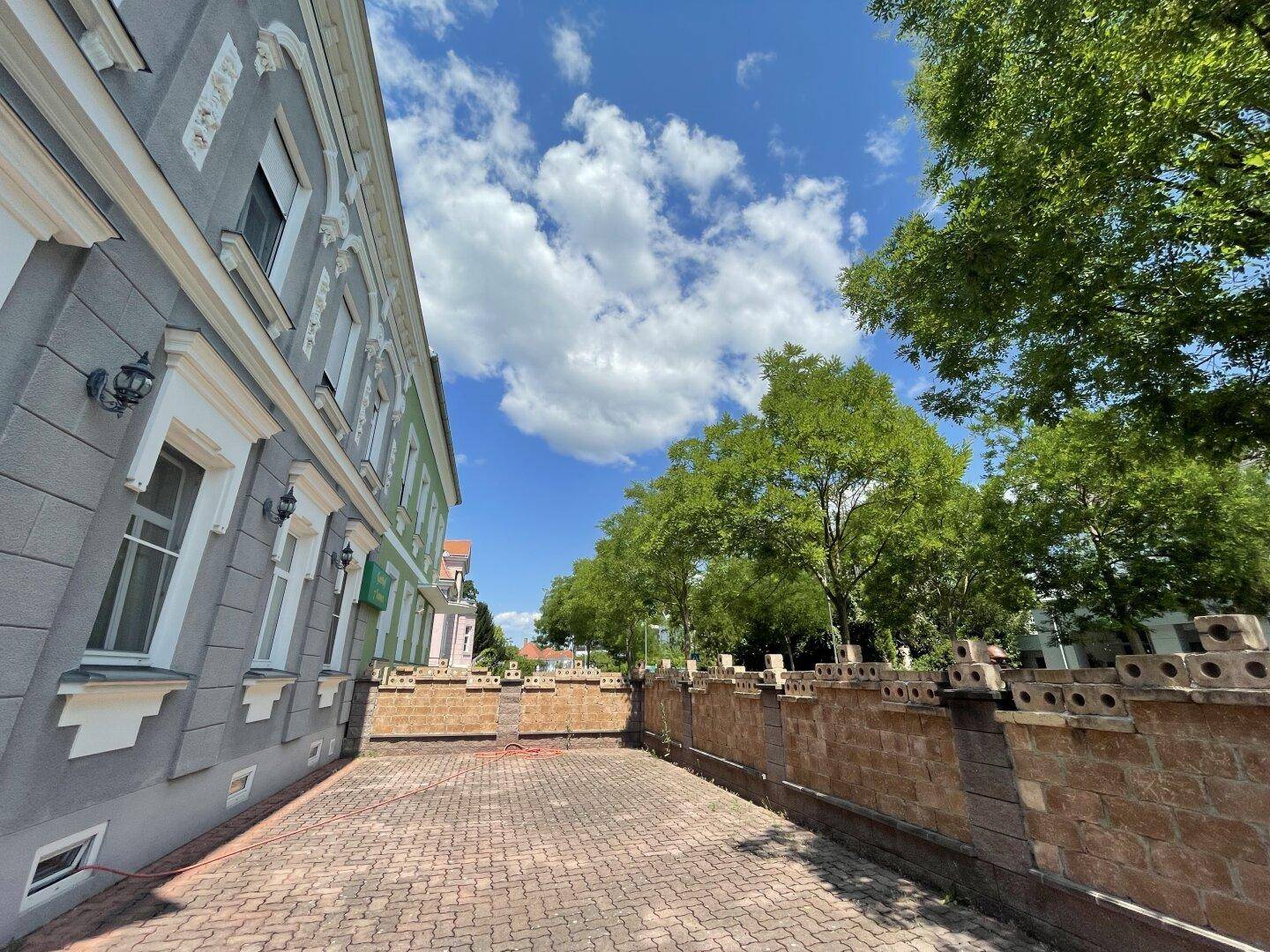 Gepflasterter Bereich neben dem historischen Gebäude mit dekorativer Mauer und Bäumen im Hintergrund.