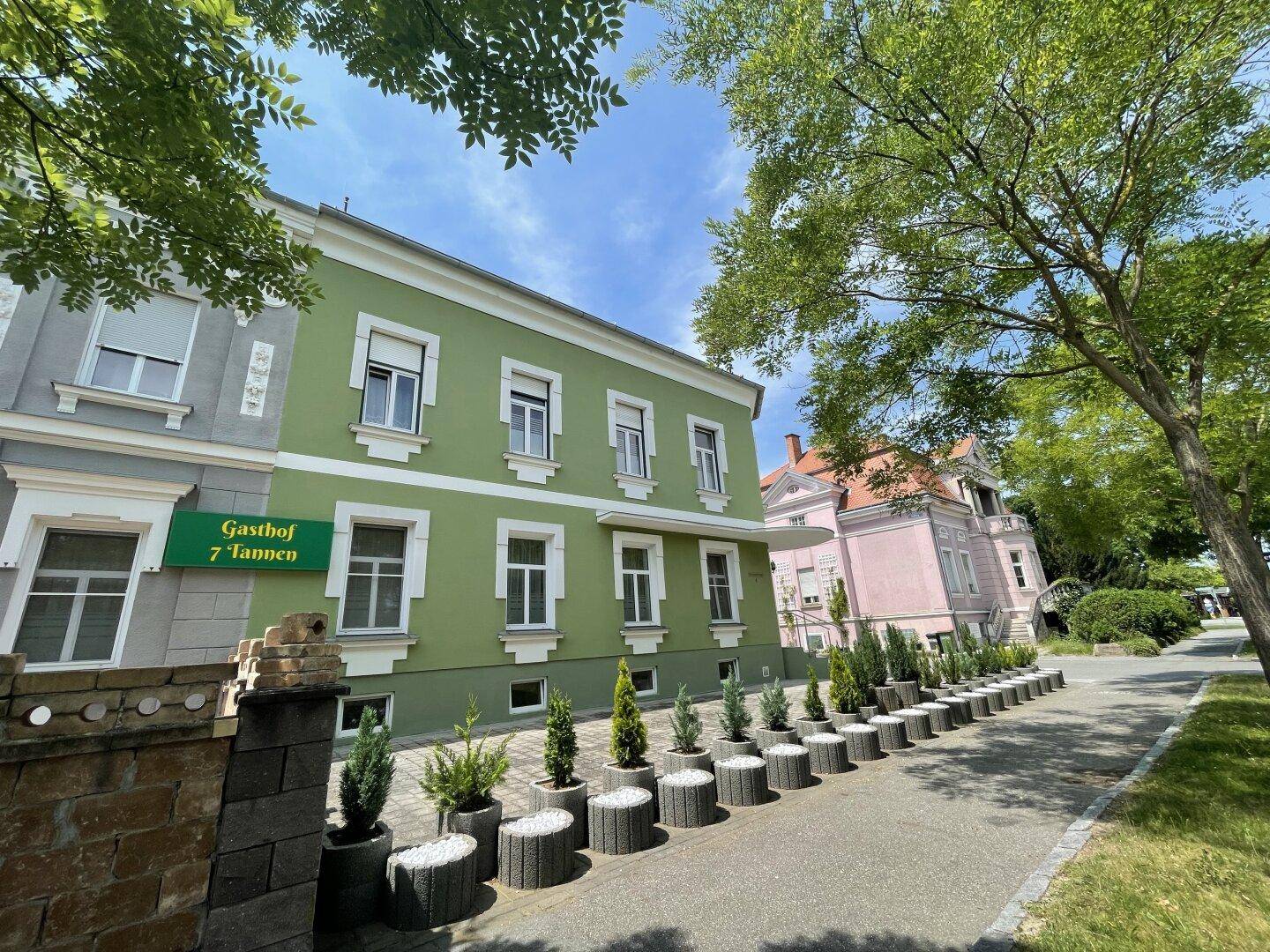 Grüne Fassade eines Gasthofs mit dekorativen Pflanzenkübeln und einem weiteren Gebäude im Hintergrund.
