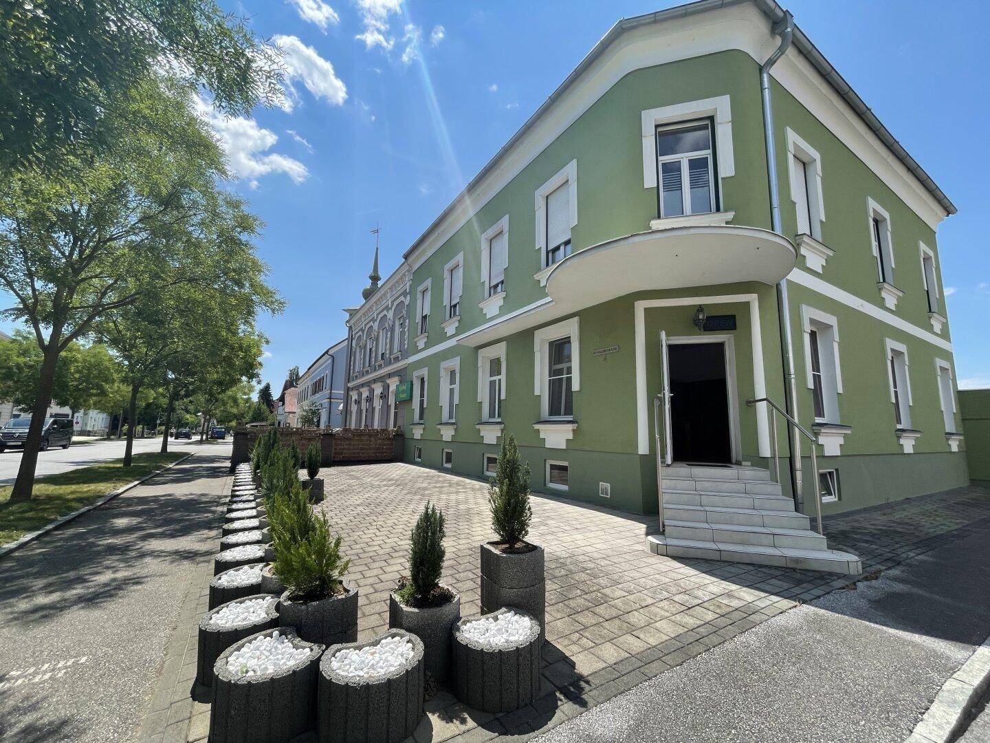 Grüne Fassade eines Gebäudes mit Eingangsbereich und gepflasterter Zufahrt unter blauem Himmel.
