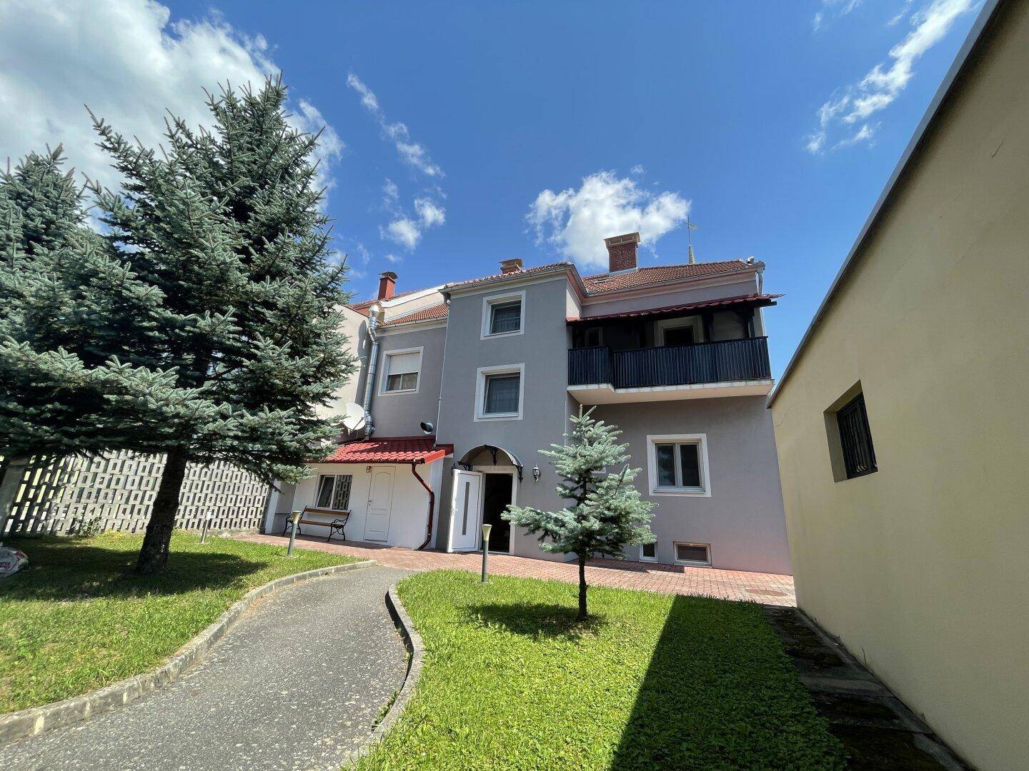 Rückseite des Gebäudes mit einem Balkon, umgeben von Rasen und Nadelbäumen.
