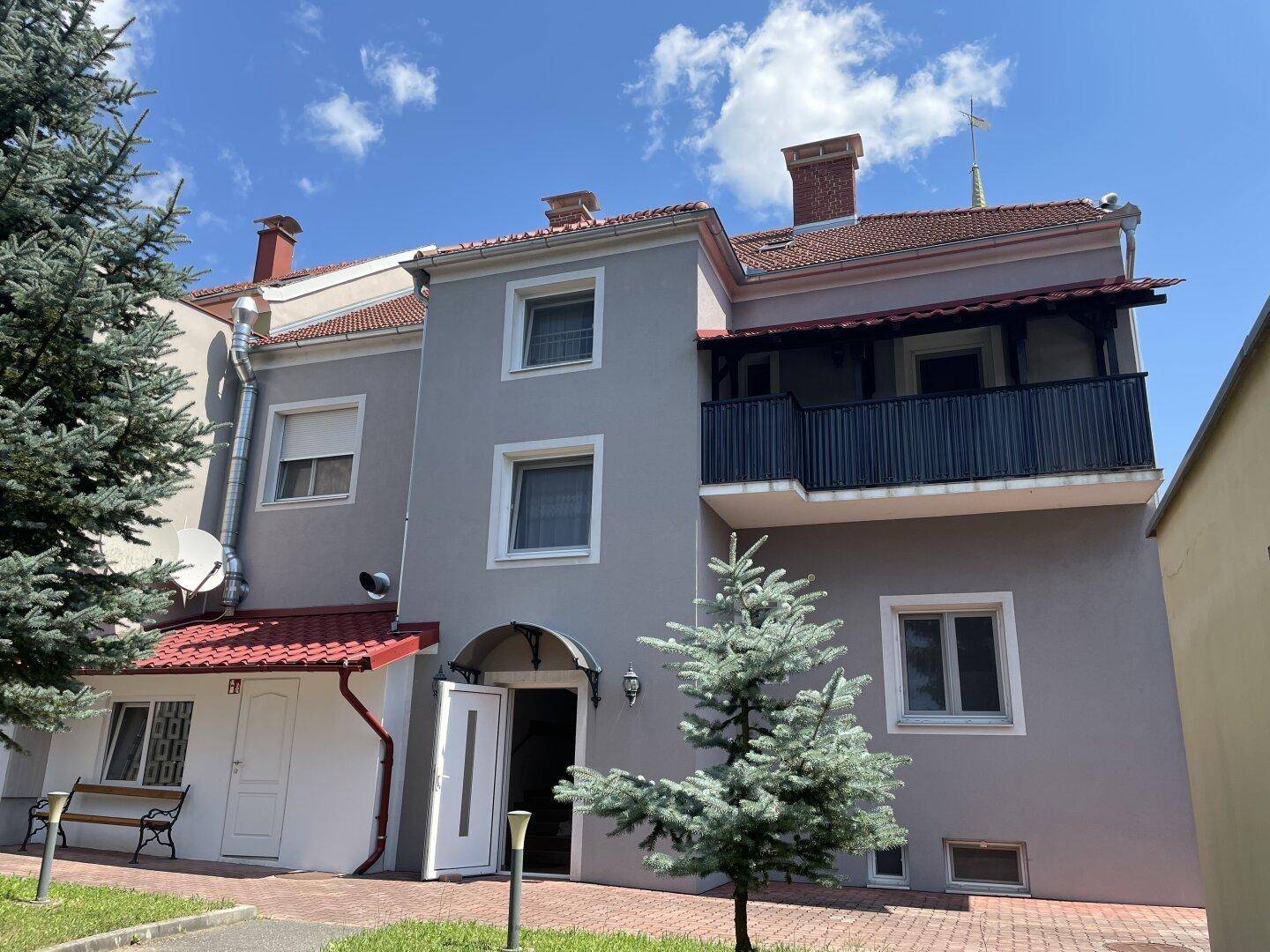 Rückseite des Gebäudes mit einem Balkon, Eingangsbereich und umgebenden Nadelbäumen.
