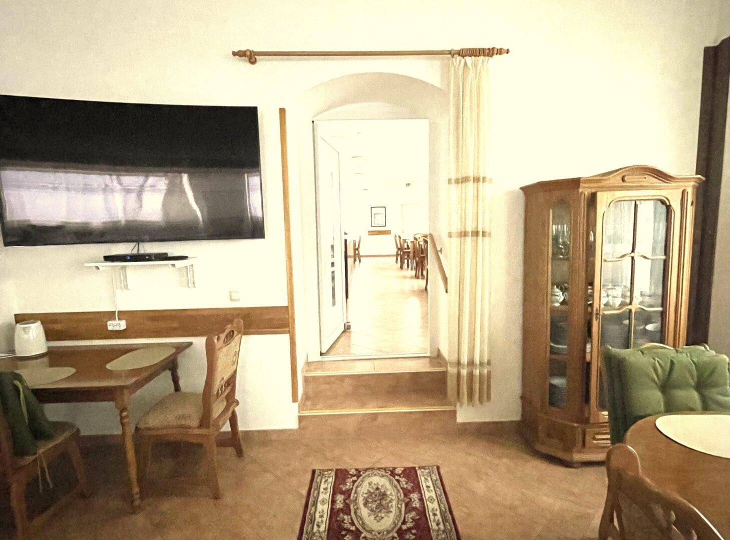 Essbereich mit Fernseher, Holztisch und Vitrine, der zu einem weiteren hellen Raum führt.