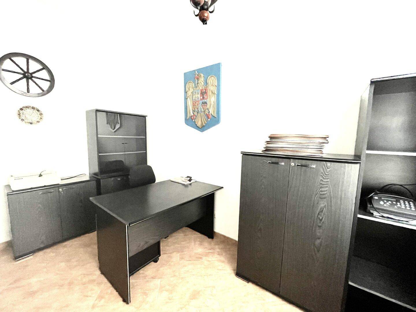 Schlichtes Büro mit dunklem Schreibtisch, Regalen und einem traditionellen Wappen an der Wand.