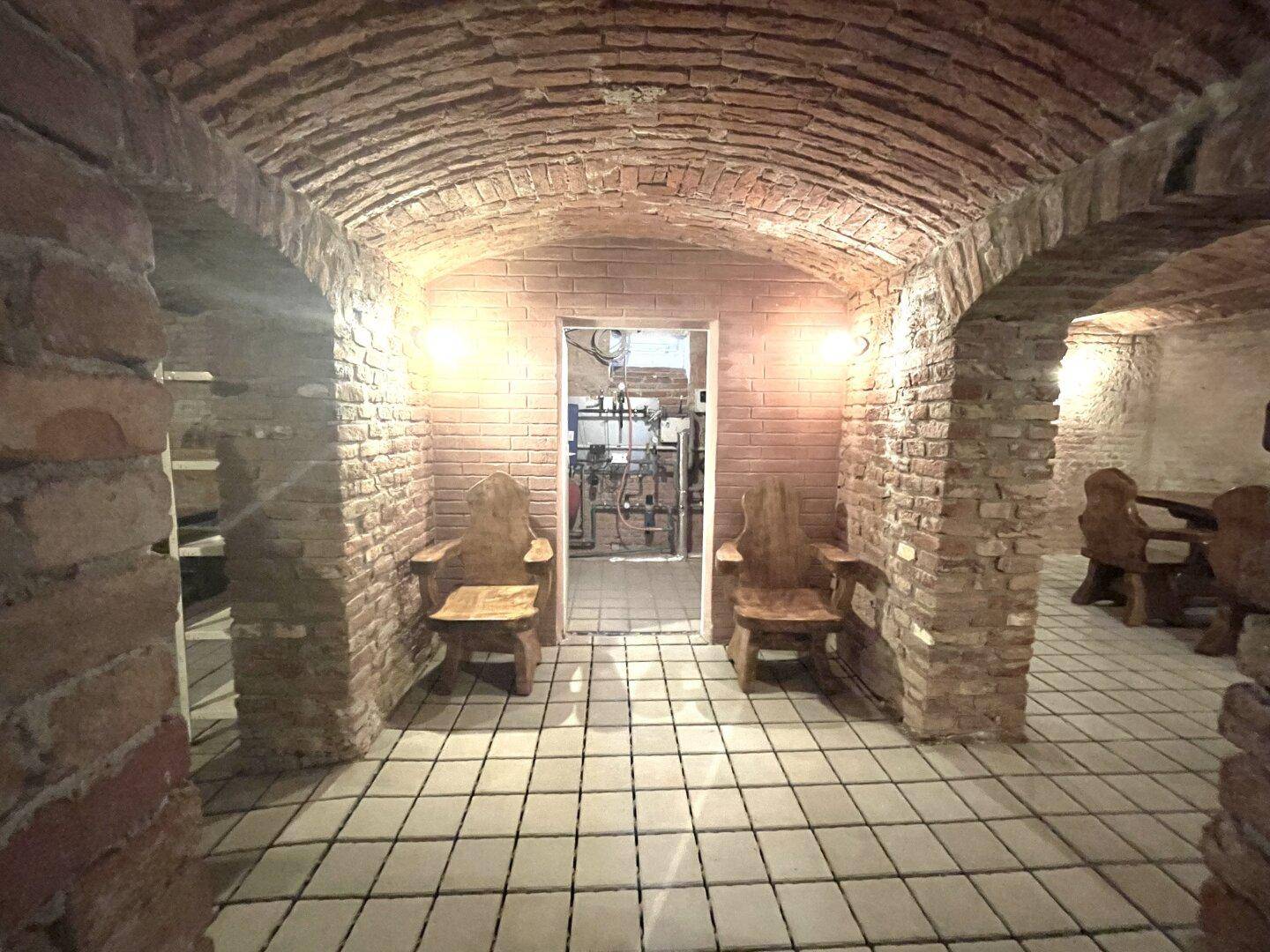 Historischer Kellerraum mit Ziegelgewölbe, Fliesenboden und rustikalen Holzstühlen.