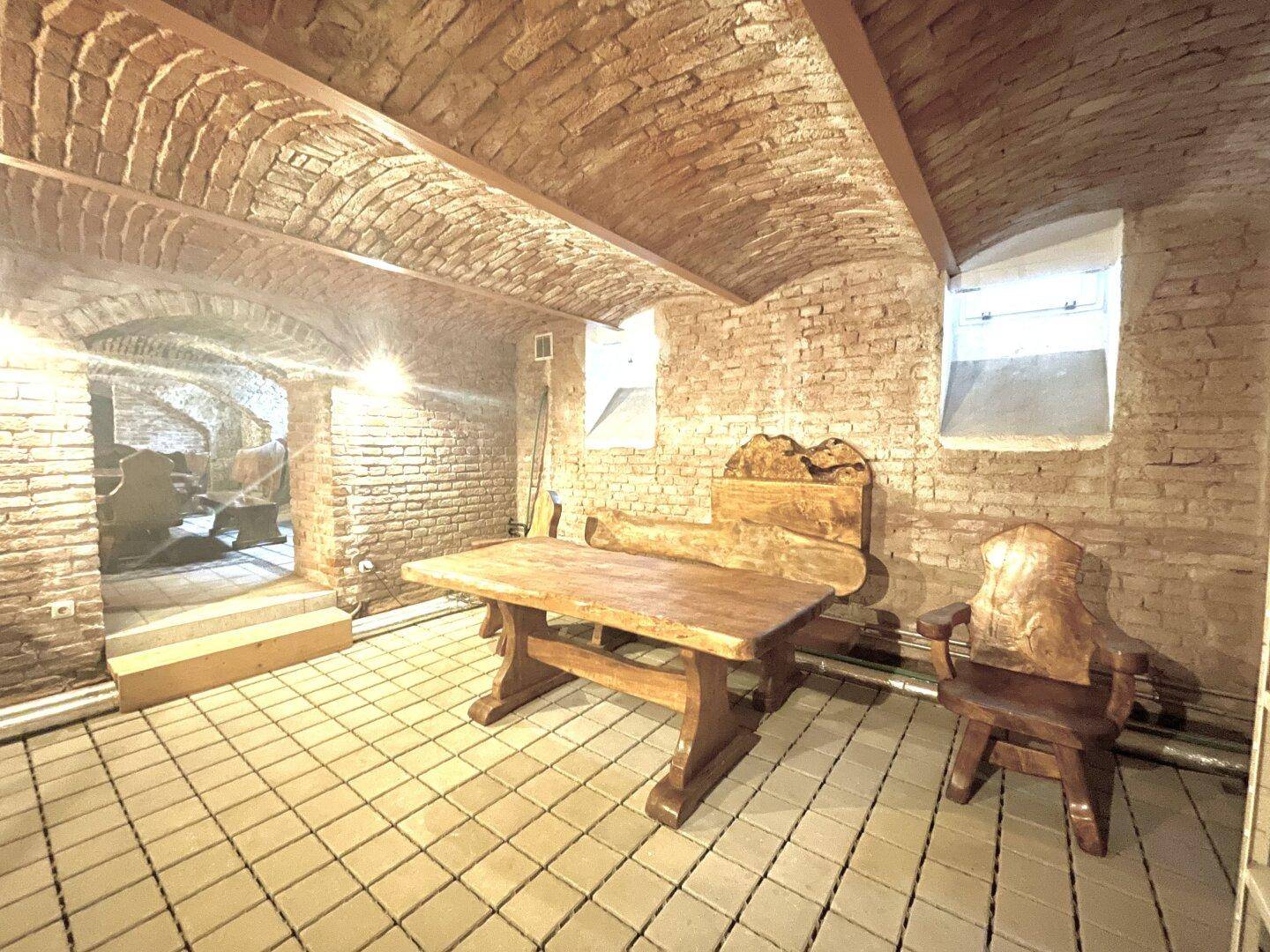 Rustikaler Kellerraum mit Ziegelgewölbe, Holztisch und Stühlen, beleuchtet von Wandlampen.