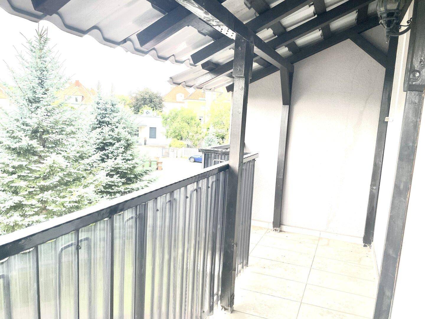 Überdachter Balkon mit Fliesenboden und Blick ins Grüne, umgeben von Bäumen.