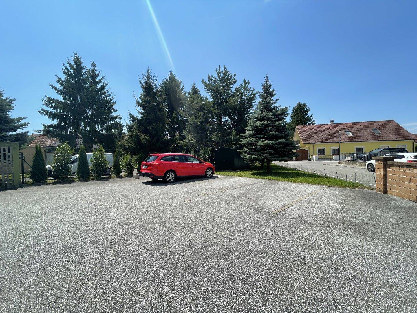 Großer Parkplatz mit einem roten Auto, umgeben von Bäumen und einem Gebäude im Hintergrund.