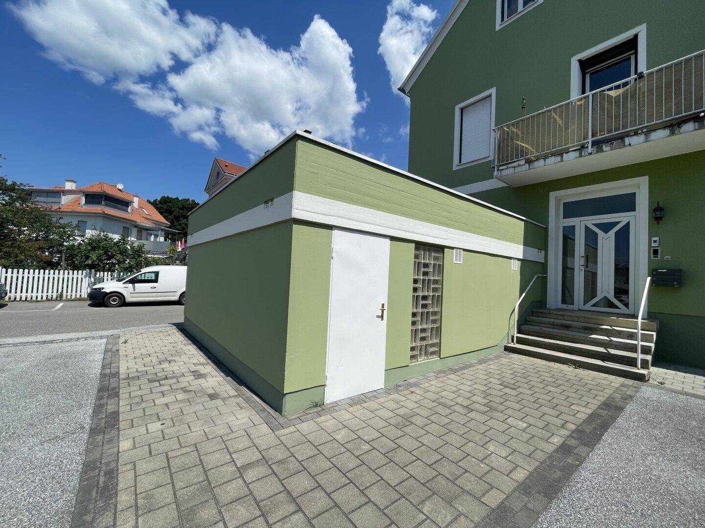 Grünes Gebäude mit Nebengebäude und gepflastertem Vorplatz, unter blauem Himmel.