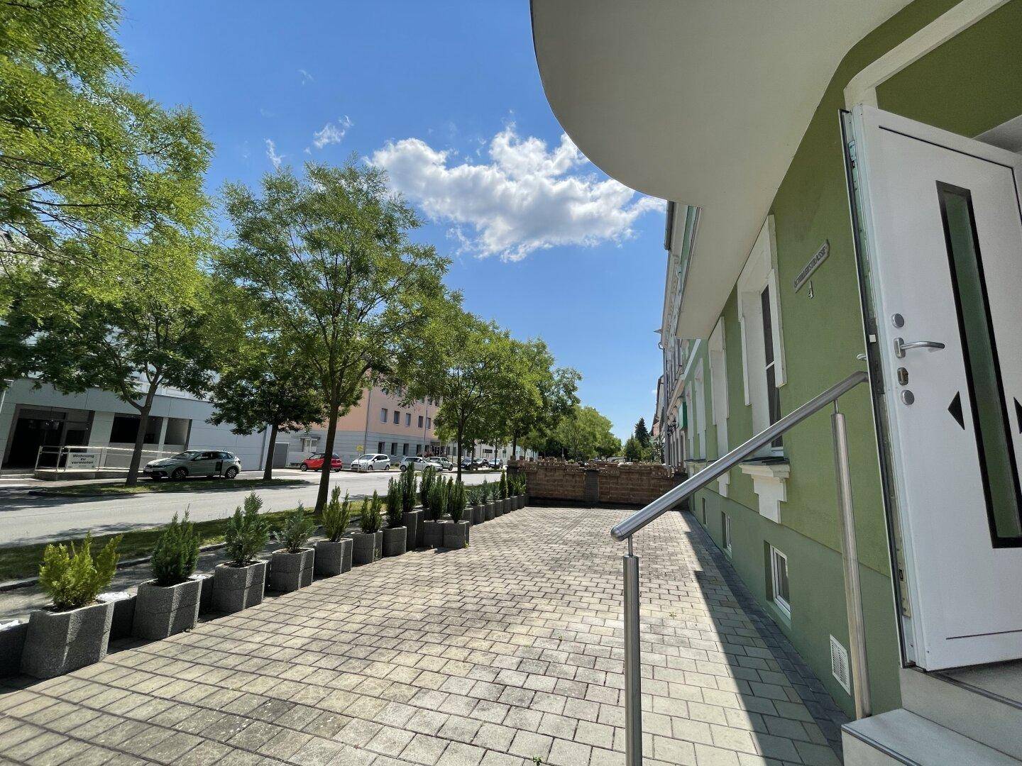 Eingangsbereich eines Gebäudes mit gepflastertem Vorplatz und dekorativen Pflanzenkübeln.
