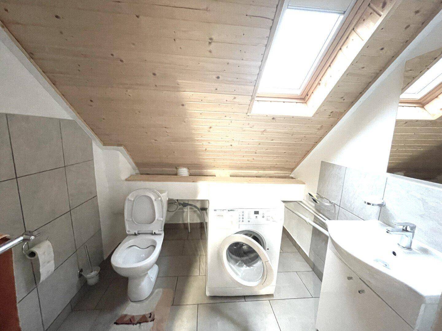 Dachgeschoss-Badezimmer mit Toilette, Waschmaschine und Waschbecken, beleuchtet durch Dachfenster.