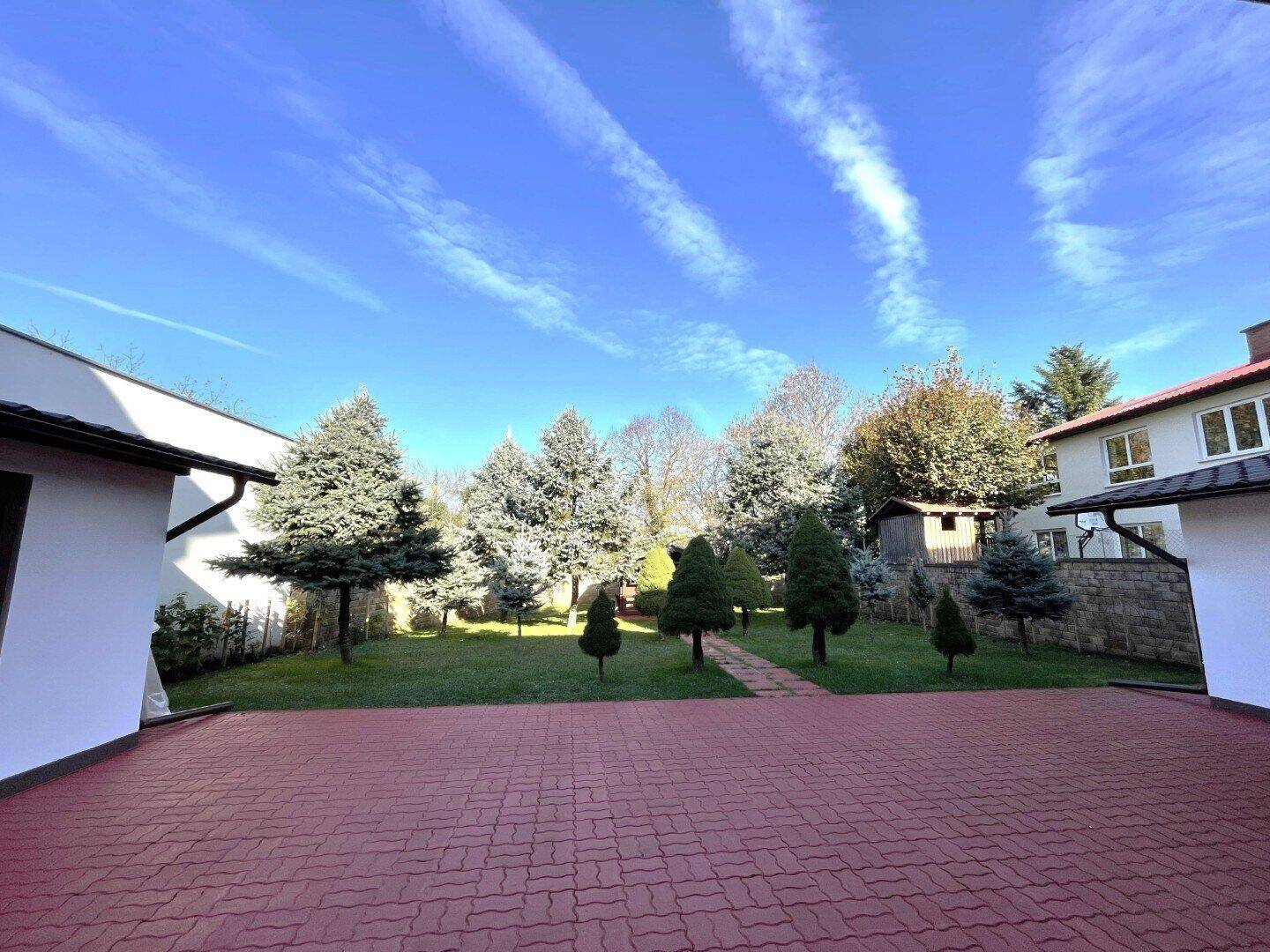 Weitläufiger Gartenbereich mit rotem Pflasterweg, grüner Rasenfläche und verschiedenen Bäumen.