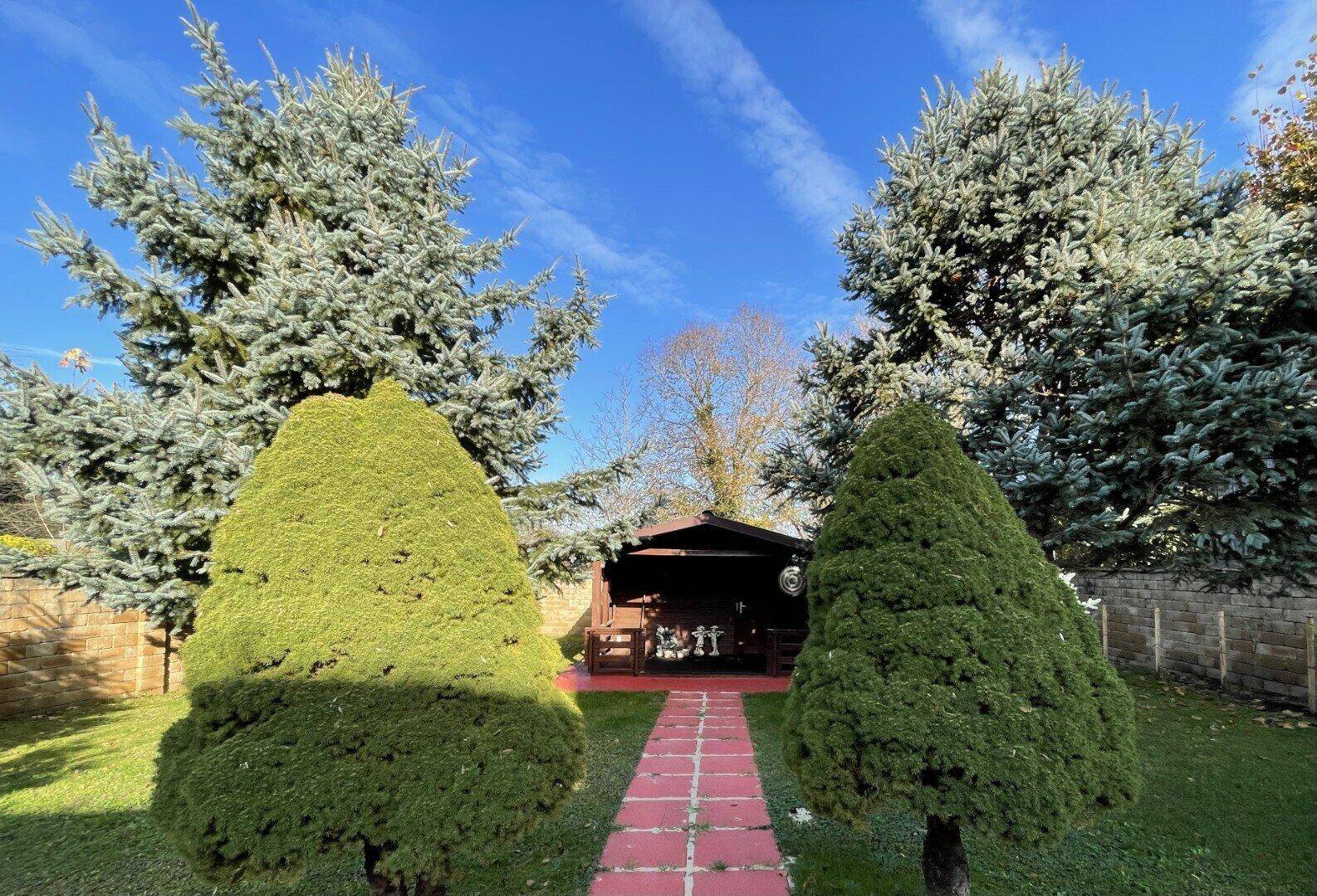 Idyllischer Gartenbereich mit einem kleinen Holzpavillon und gepflegten Nadelbäumen, sonnig und grün.