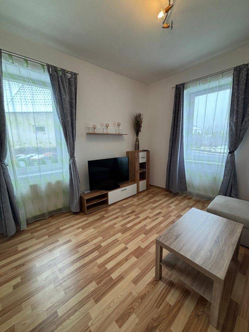 Wohnzimmer mit Laminatboden, Fernseher und Vorhängen an den Fenstern.
