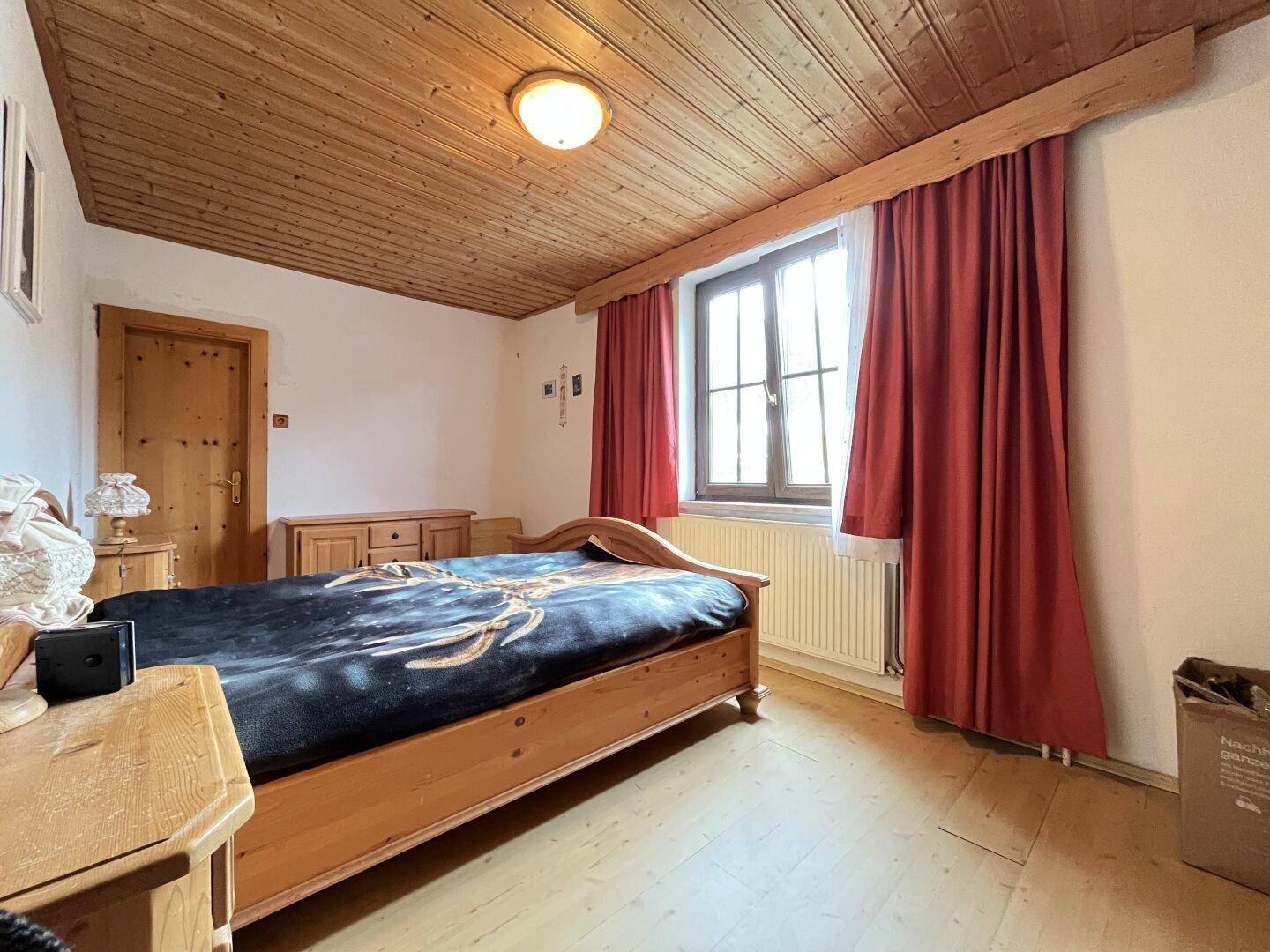 Schlafzimmer mit Holzbett, Laminatboden und roten Vorhängen am Fenster.