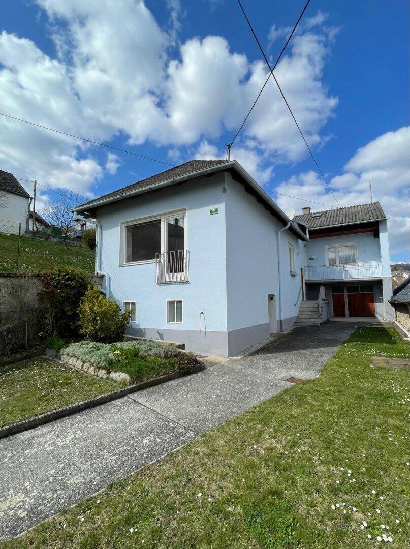 Seitenansicht eines hellblauen Hauses mit Auffahrt und Hanglage im ländlichen Umfeld.