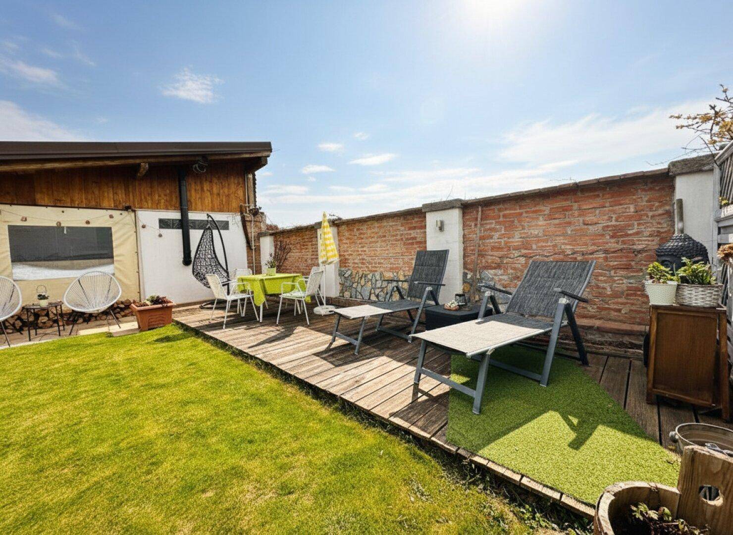 Sonniger Gartenbereich mit Rasen, Holzterrasse und Liegestühlen für entspannte Stunden im Freien.