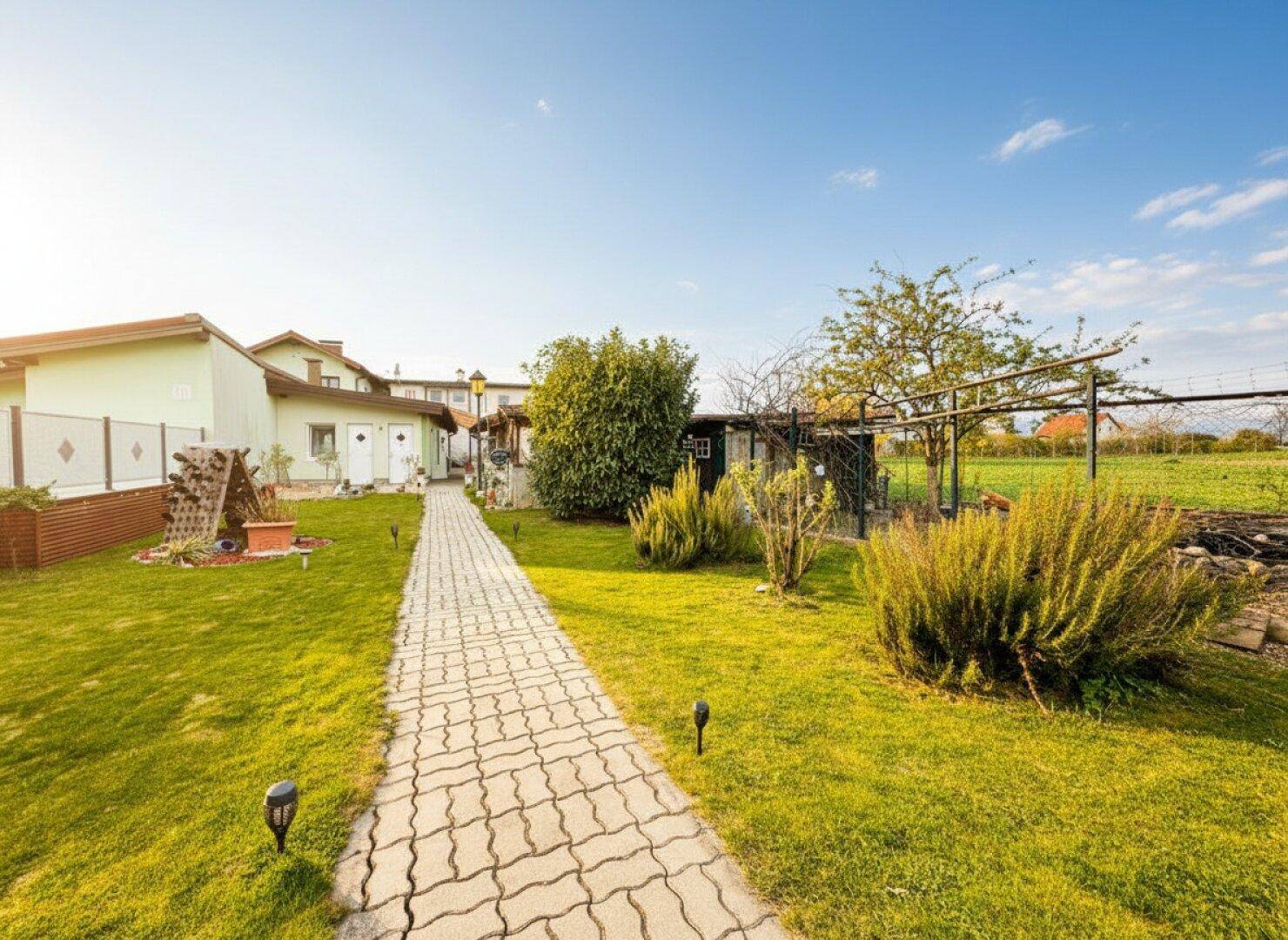 Langer gepflasterter Weg durch einen grünen Garten mit Büschen und Gebäuden im Hintergrund unter blauem Himmel.