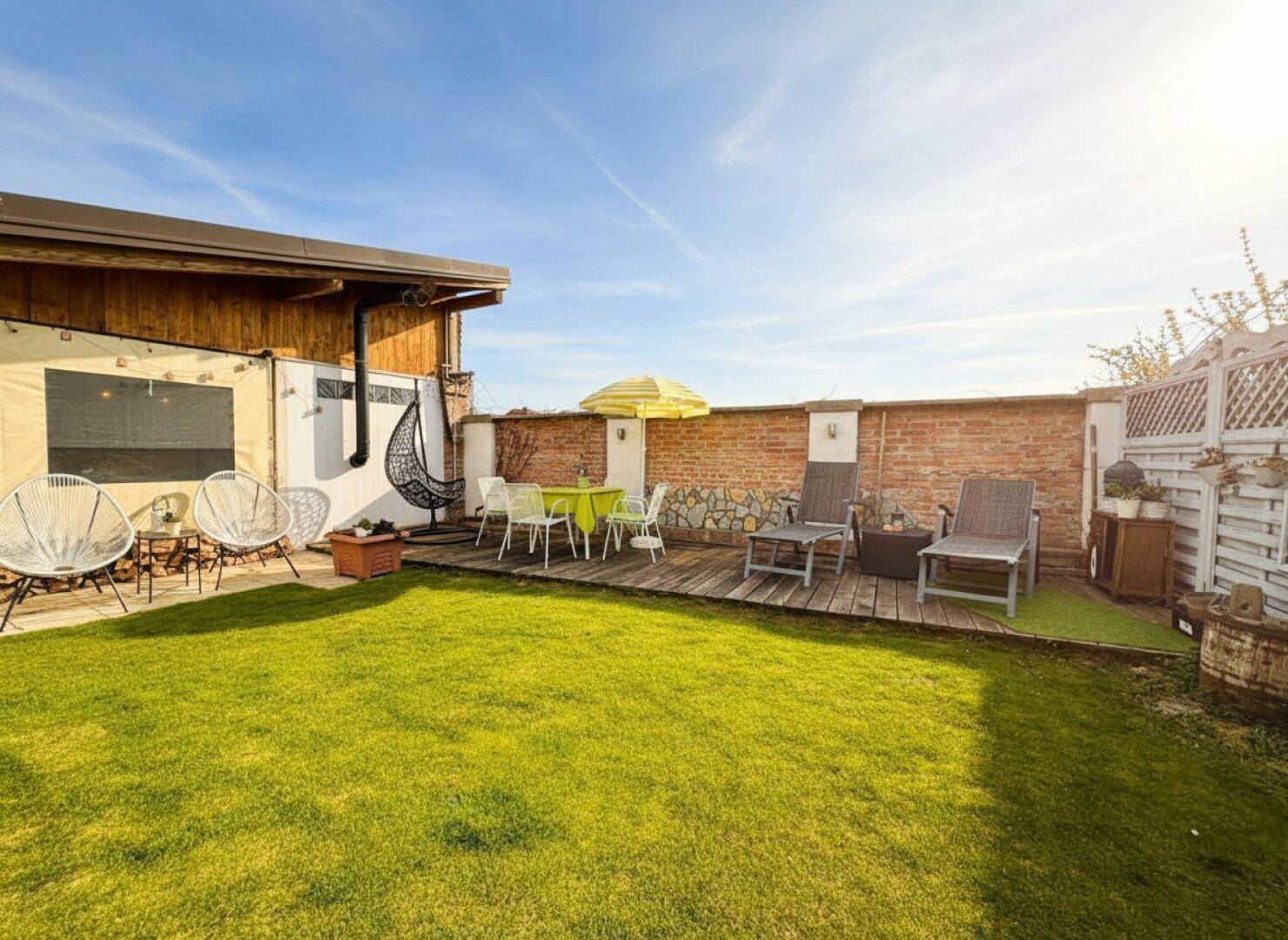 Sonniger Gartenbereich mit Rasen, Holzterrasse und verschiedenen Sitzgelegenheiten für Entspannung im Freien.