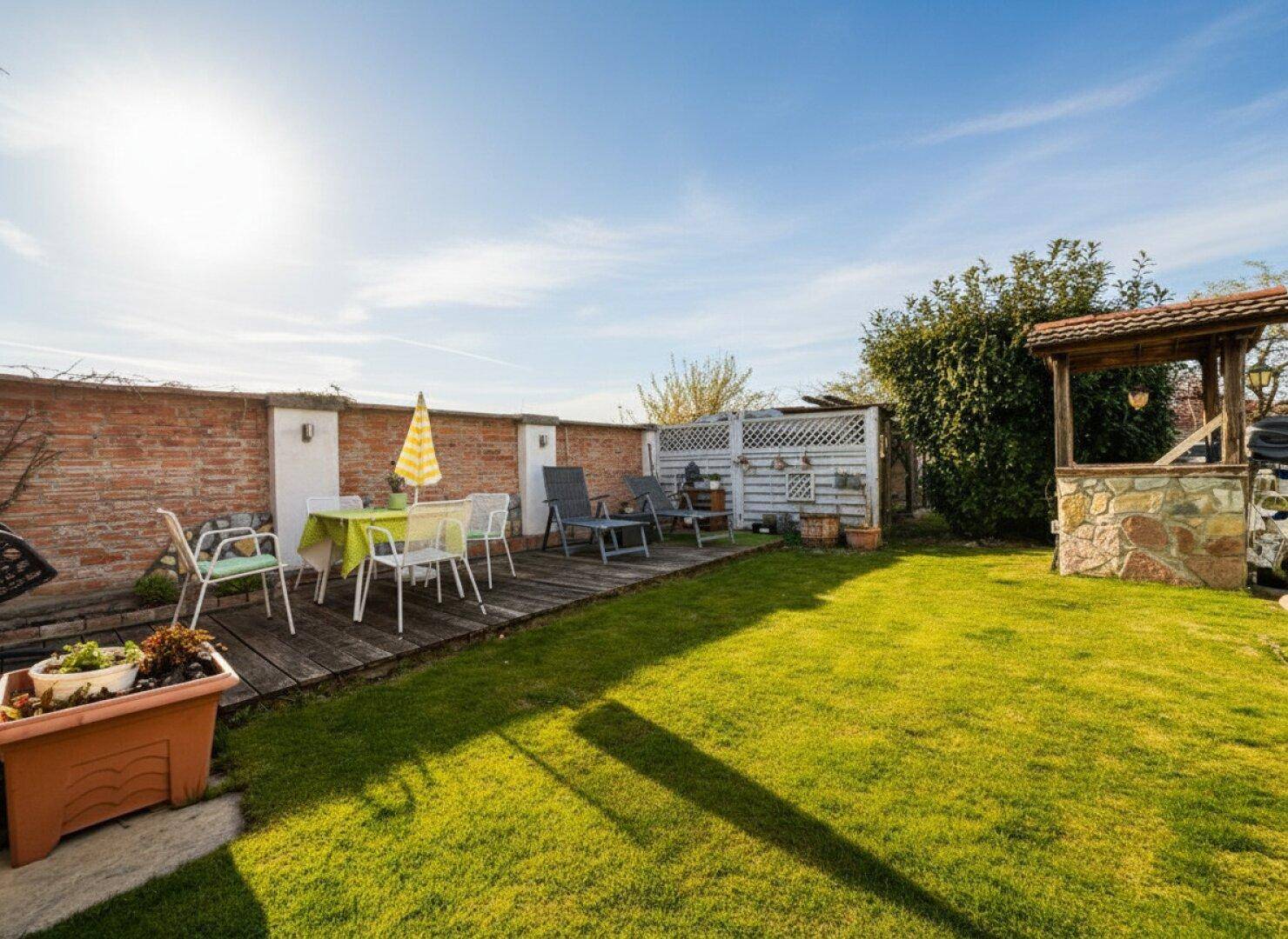 Sonniger Garten mit Rasenfläche, Holzterrasse und einem Brunnen, umgeben von einer Ziegelmauer.