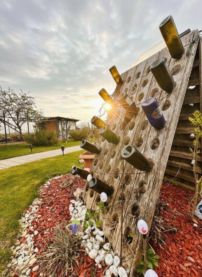 Kreative Gartendekoration mit Weinflaschen in einem Holzgestell, umgeben von Kies und Pflanzen.