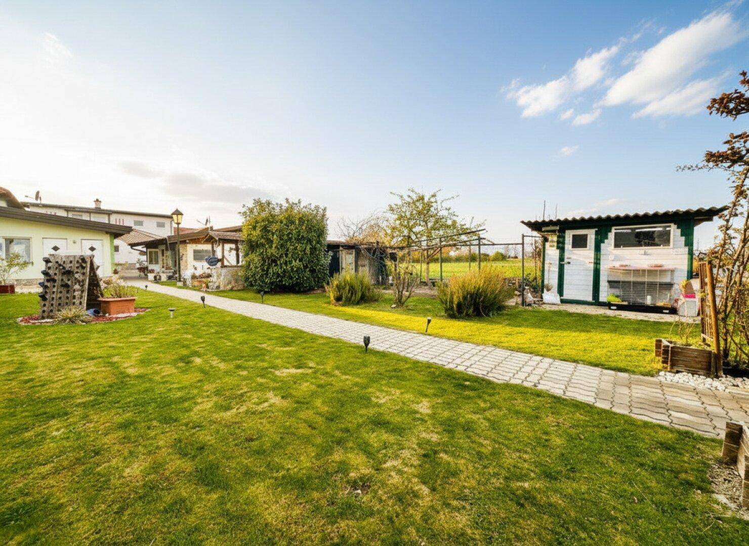 Weitläufiger Gartenbereich mit gepflastertem Weg, Rasenflächen und verschiedenen Nebengebäuden.