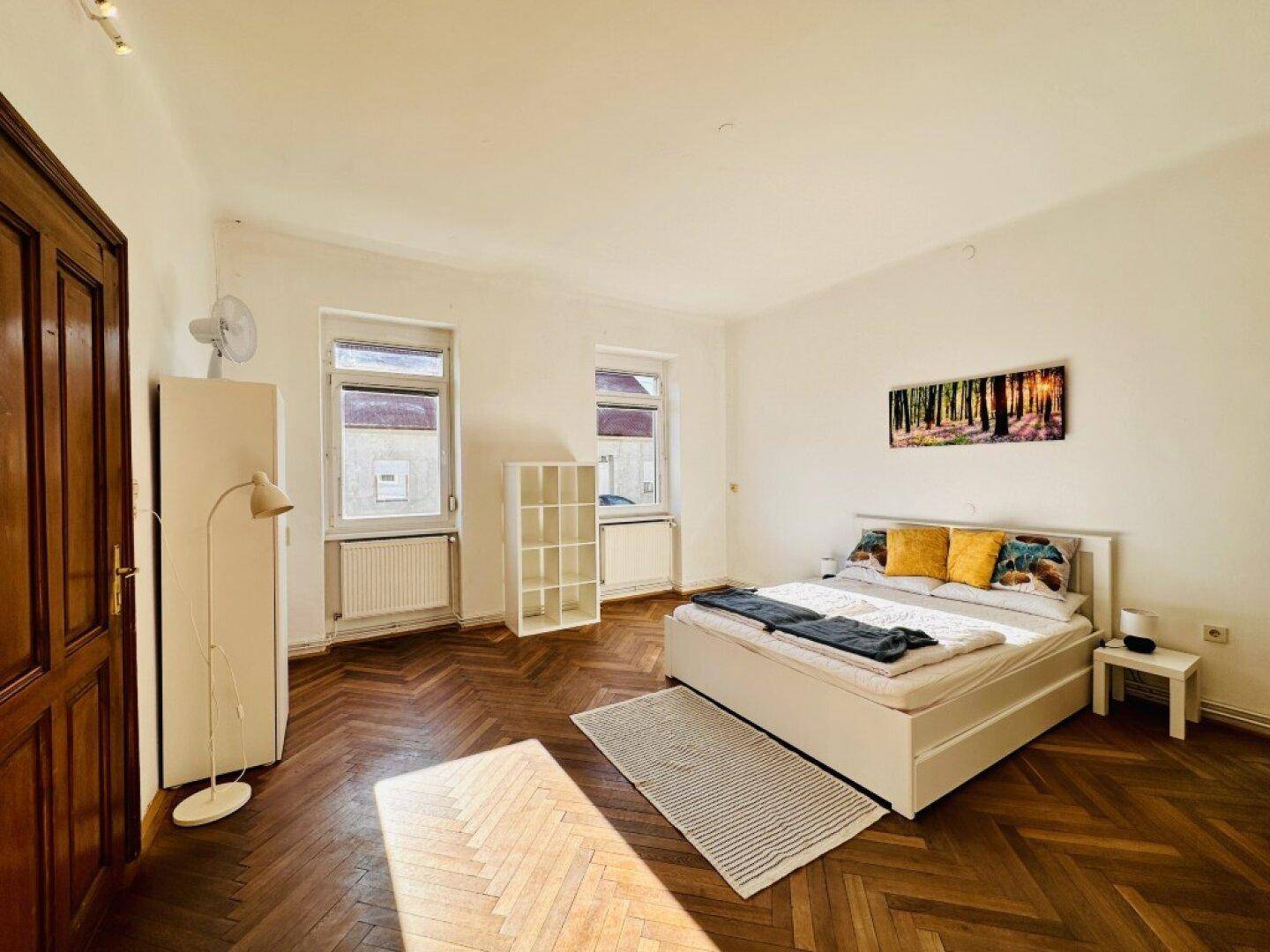 Helles Schlafzimmer mit Fischgrätparkett, einem Doppelbett und zwei Fenstern.