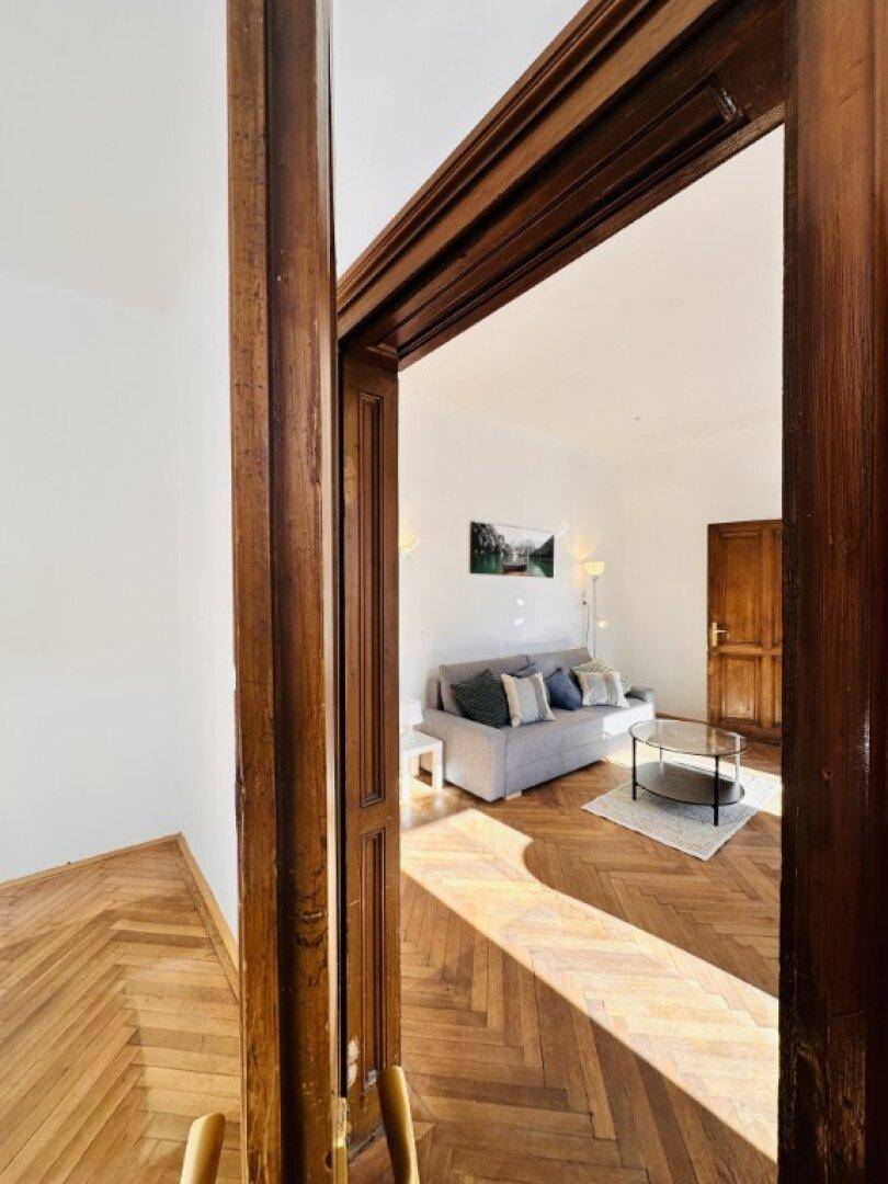 Blick durch eine Holztür in ein helles Wohnzimmer mit Fischgrätparkett und Sofa.
