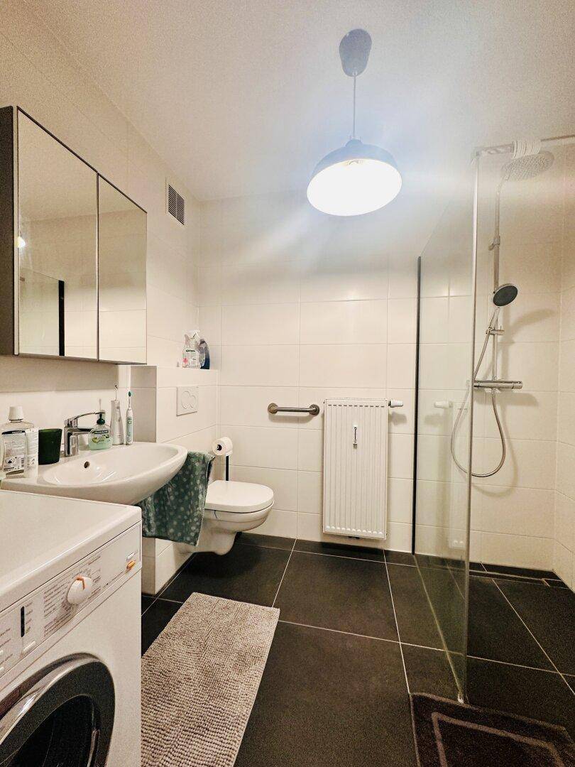 Modernes Badezimmer mit ebenerdiger Dusche, Waschbecken und Waschmaschine, dunkel gefliest.