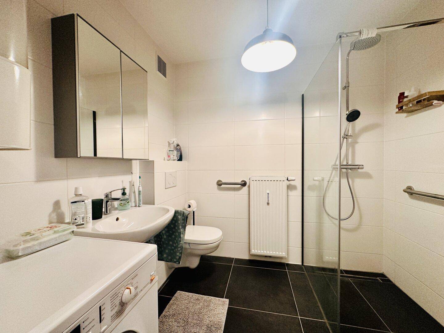 Helles Badezimmer mit Dusche, Waschbecken, WC und Waschmaschine, ausgestattet mit Spiegelschrank.