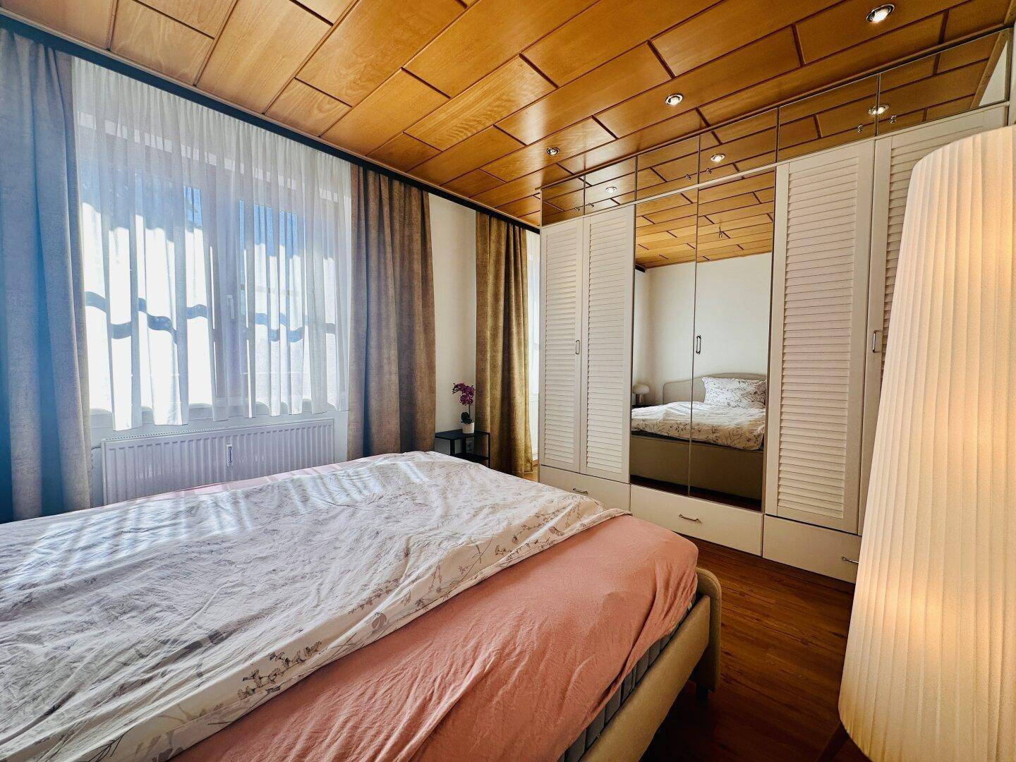Schlafzimmer mit Holzboden, Holzdecke und großem Spiegelschrank, hell durch Fenster.