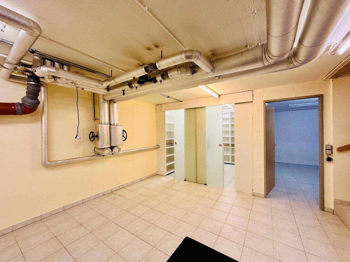Kellerraum mit sichtbaren Rohren an der Decke und gefliestem Boden, ideal als Abstellraum.